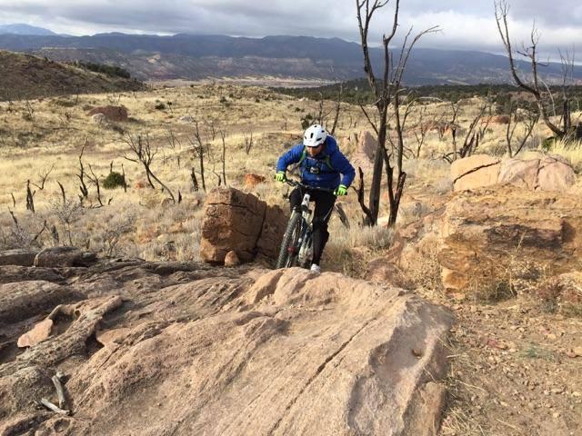 A mountain biker navigates a rocky trail, surrounded by dry grass and sparse trees, with distant mountains under a cloudy sky. Oil Well Flats mountain bike trail.