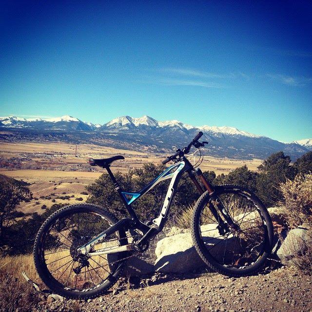 A mountain bike resting on a rock with a scenic view of snow-capped mountains in the background, under a clear blue sky. The expansive landscape features rolling hills and a valley, showcasing the beauty of outdoor adventure and cycling. Arkansas Hills mountain bike trail.