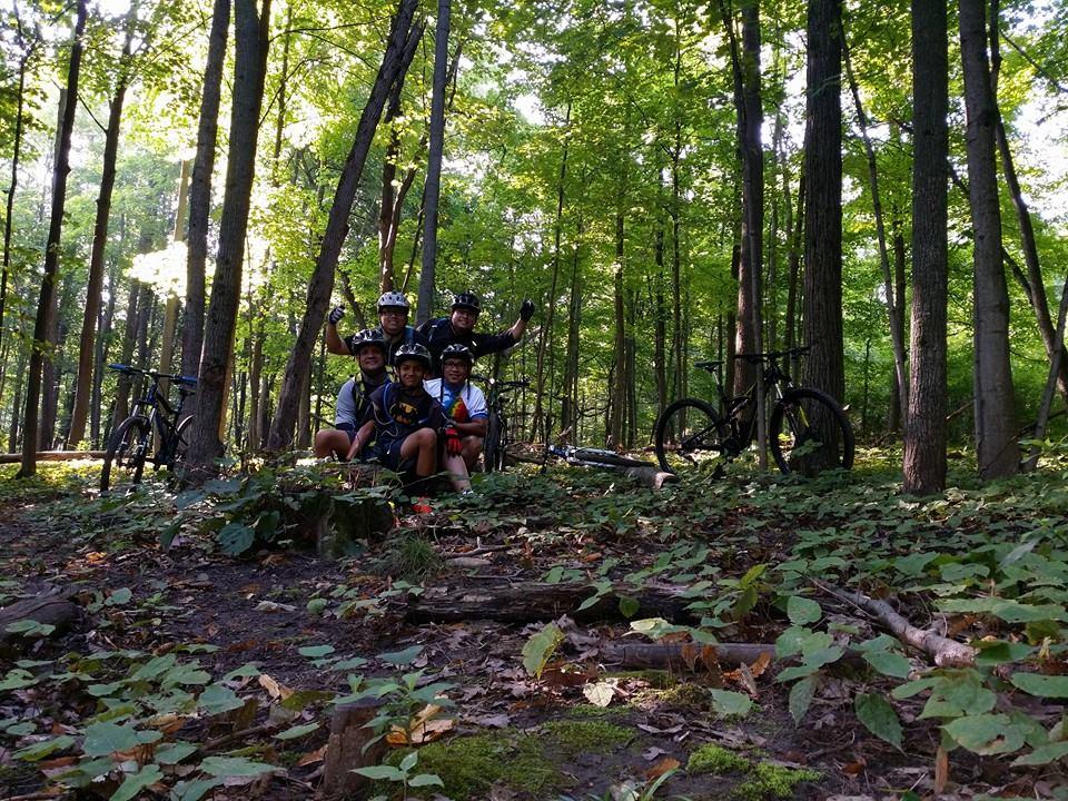 Group of five people posing for a photo in a forested area, surrounded by tall trees and greenery. They are wearing helmets and cycling gear, with mountain bikes parked nearby. The scene is bright and vibrant, suggesting a fun outdoor biking adventure. Raceway Woods mountain bike trail.