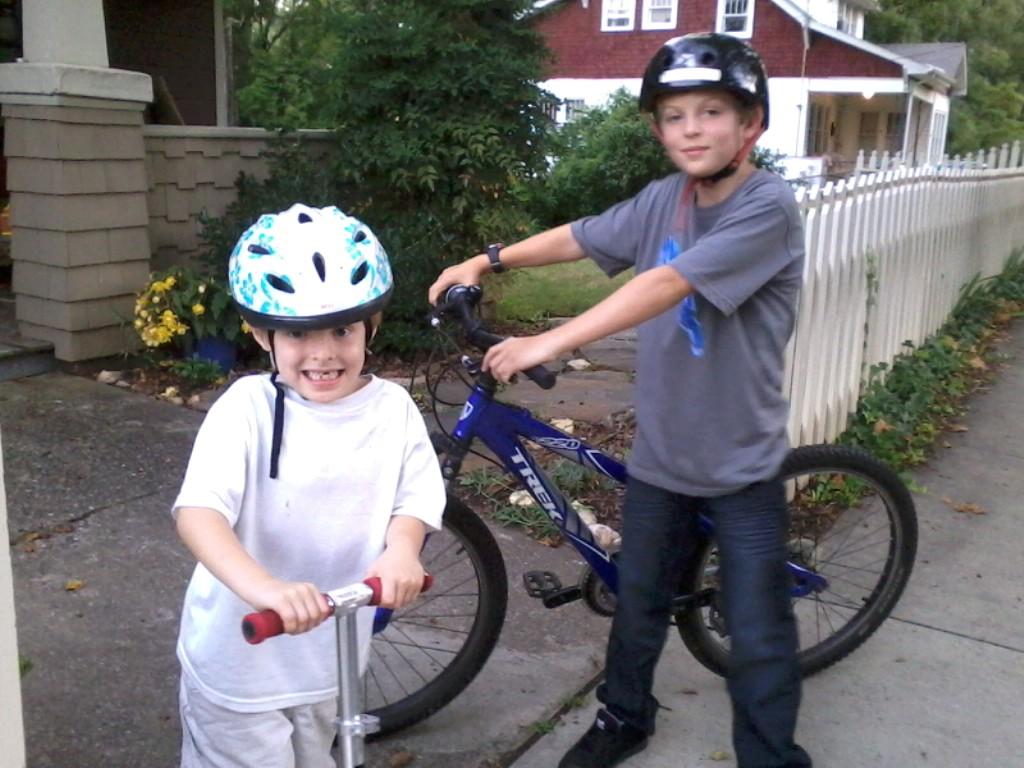 Trek 8000: Two children are playing outside. One child, wearing a blue and white helmet, is smiling and standing next to a scooter. The other child, wearing a black helmet, is standing beside a blue bicycle, looking at the camera. The background features a residential area with a white picket fence and greenery.