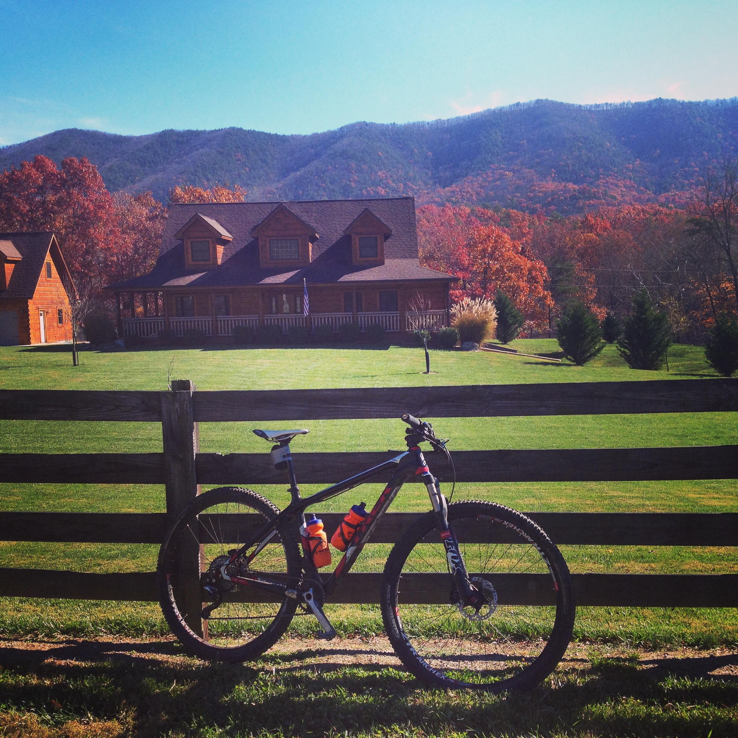 A mountain bike leans against a wooden fence in front of a large house surrounded by lush green grass and colorful fall foliage. The backdrop features distant mountains under a clear blue sky. Carvin's Cove Trail system mountain bike trail.