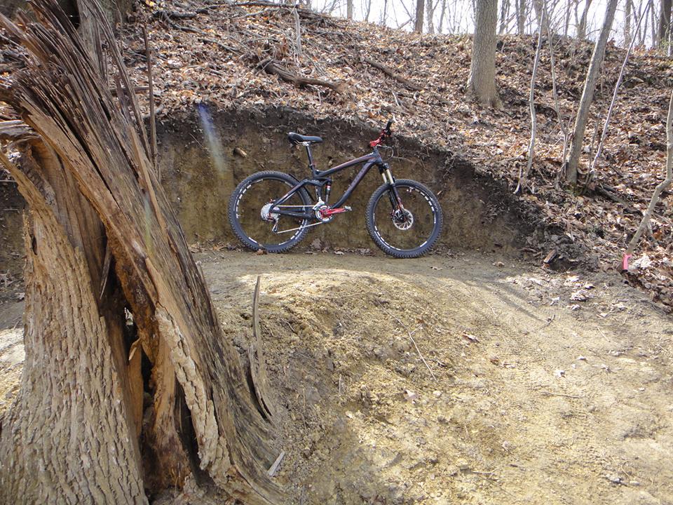 A mountain bike stands on a dirt path in a wooded area, with a backdrop of trees and bare ground covered in fallen leaves. A large, partially decayed tree trunk is prominent in the foreground, and the terrain features a dirt jump or berm, indicating a biking trail. Kickapoo mountain bike trail.