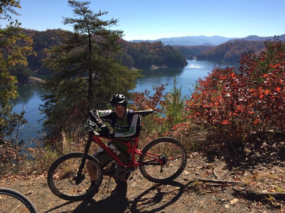 A mountain biker resting on a red bike next to a scenic overlook of a lake surrounded by trees with autumn foliage. The clear blue sky and distant mountains complete the picturesque landscape. Tsali Thompson Loop mountain bike trail.