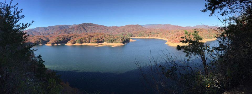 A panoramic view of a serene lake surrounded by mountains, showcasing vibrant autumn foliage in shades of red, orange, and yellow. The clear blue sky reflects on the water's surface, with trees framing the foreground and creating a tranquil natural scene. Tsali Thompson Loop mountain bike trail.