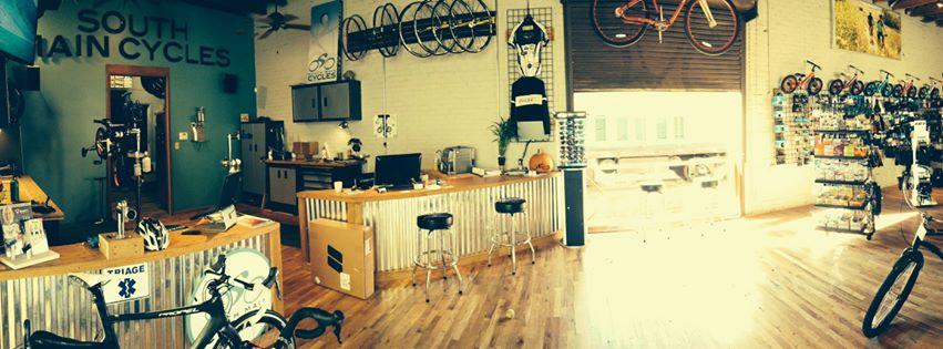 Panoramic view of a bicycle shop interior featuring a wood floor, a service counter, and display areas for bikes and accessories. The wall is decorated with bike wheels and cycling gear, while items for sale are neatly arranged on shelves. The shop is well-lit, creating a welcoming atmosphere.