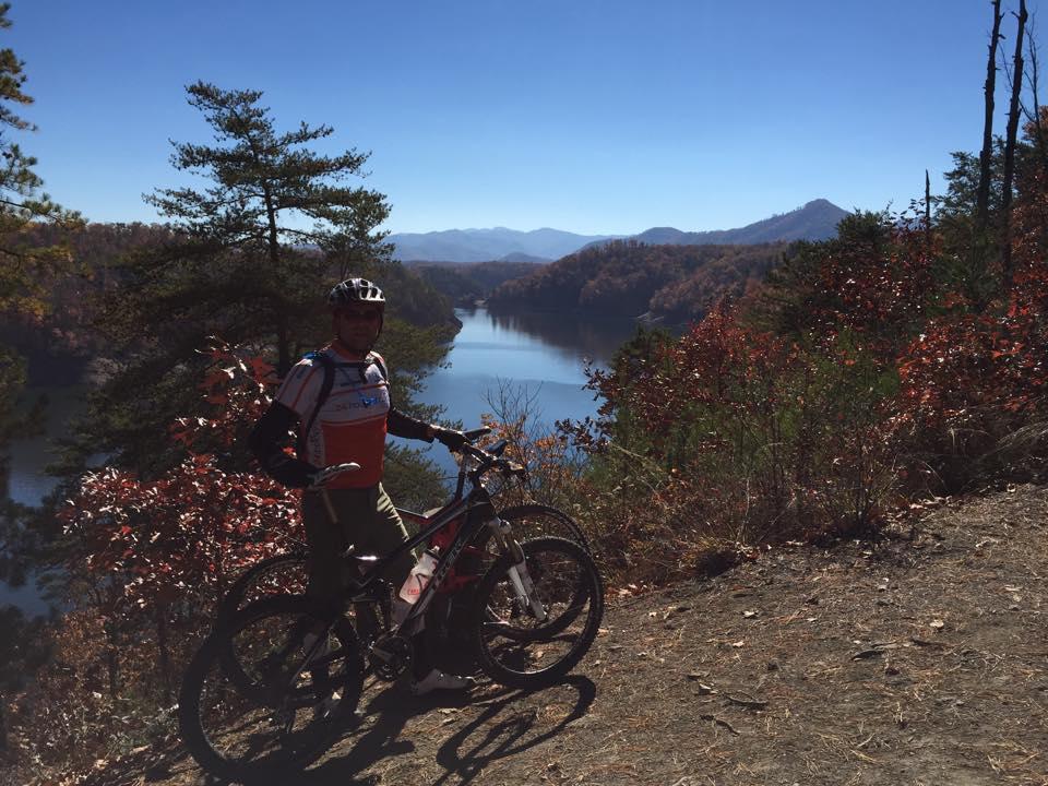 A mountain biker stands beside a bicycle on a dirt path overlooking a serene lake surrounded by autumn foliage. The background features mountains under a clear blue sky, with warm-colored leaves creating a picturesque fall scene. Tsali Thompson Loop mountain bike trail.