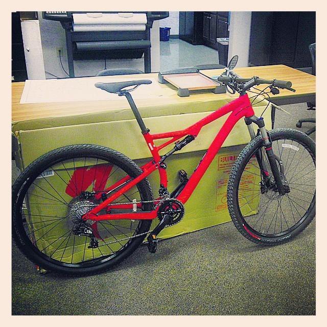 Specialized Epic: A bright red mountain bike leaning against a large cardboard box on a carpeted surface. In the background, there are office furnishings, including a large printer and wooden tables. The bike features thick tires and a modern frame design, suggesting it's ready for both outdoor trails and casual rides.