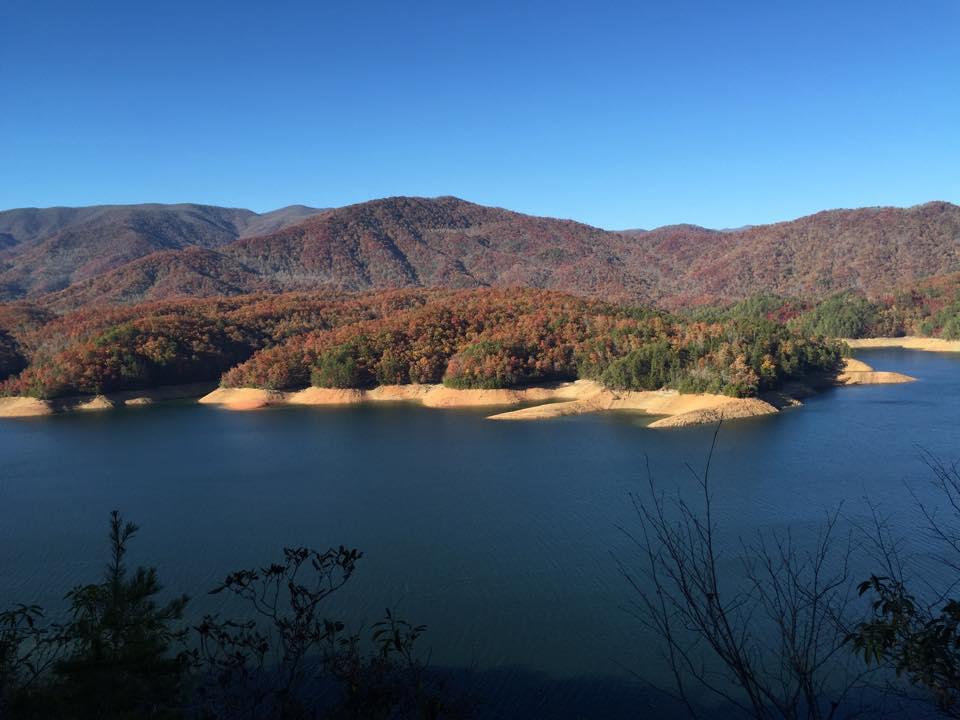 A panoramic view of a tranquil lake surrounded by vibrant autumn foliage, with rolling mountains in the background under a clear blue sky. The shoreline features sandy banks and patches of greenery, reflecting the warm, colorful hues of fall. Tsali Thompson Loop mountain bike trail.