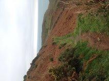A scenic view of a hillside with reddish-brown terrain and patches of green vegetation, overlooking the ocean in the distance under a cloudy sky. Sorel Point to L'Etacq mountain bike trail.