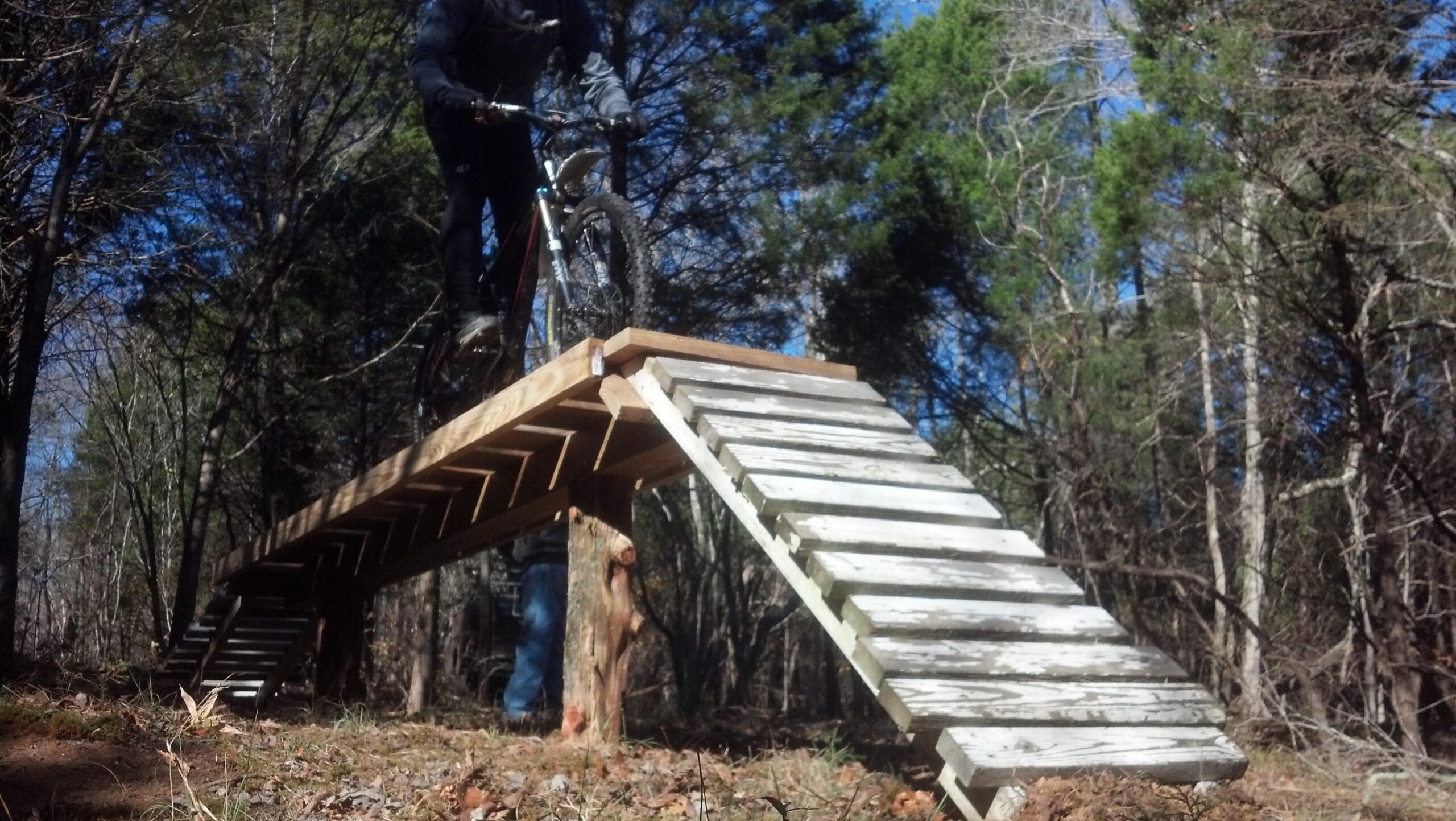 A mountain biker riding a wooden ramp in a forested area, showcasing an elevated platform leading to a sloped descent. The surroundings are filled with trees and underbrush, and the sky is clear. The biker appears focused and balanced as they navigate the ramp. Green River mountain bike trail.