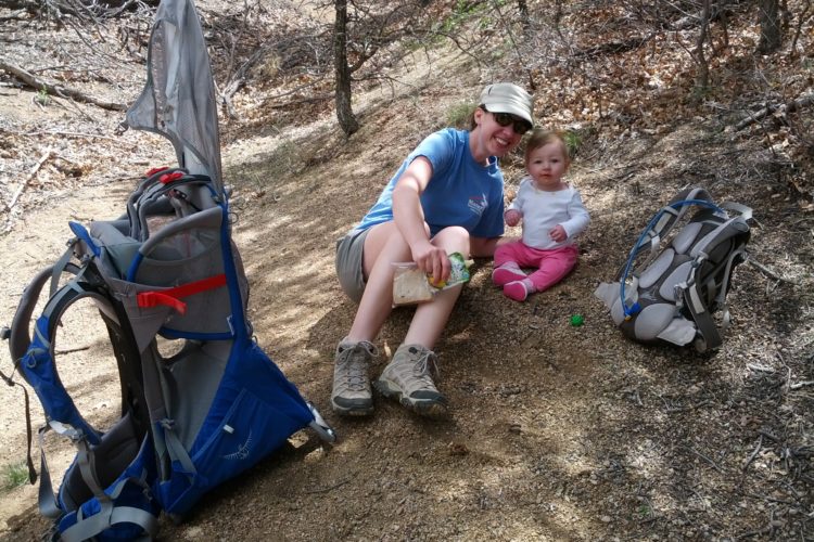 A woman sitting on a trail with a toddler beside her, both enjoying a break during a hike. The woman is smiling and holding a snack, while the child, dressed in pink, is sitting on the ground and looking curiously at the camera. Two backpacks, one blue and one gray, are positioned nearby on the gravel path, surrounded by trees and nature.