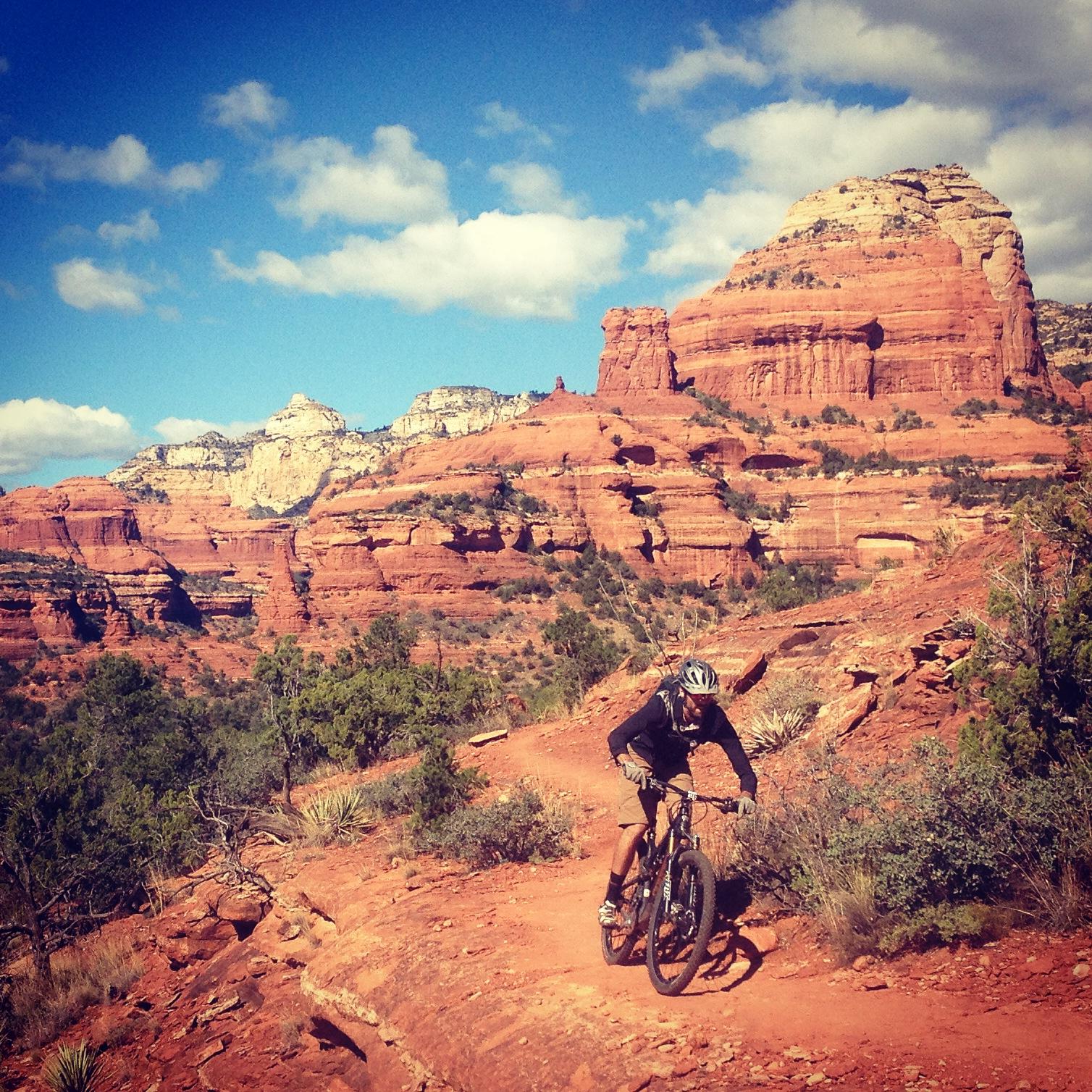 A mountain biker navigating a rocky trail in a desert landscape, surrounded by red rock formations and blue skies with scattered clouds. Upper Dry Creek Area Trails mountain bike trail.