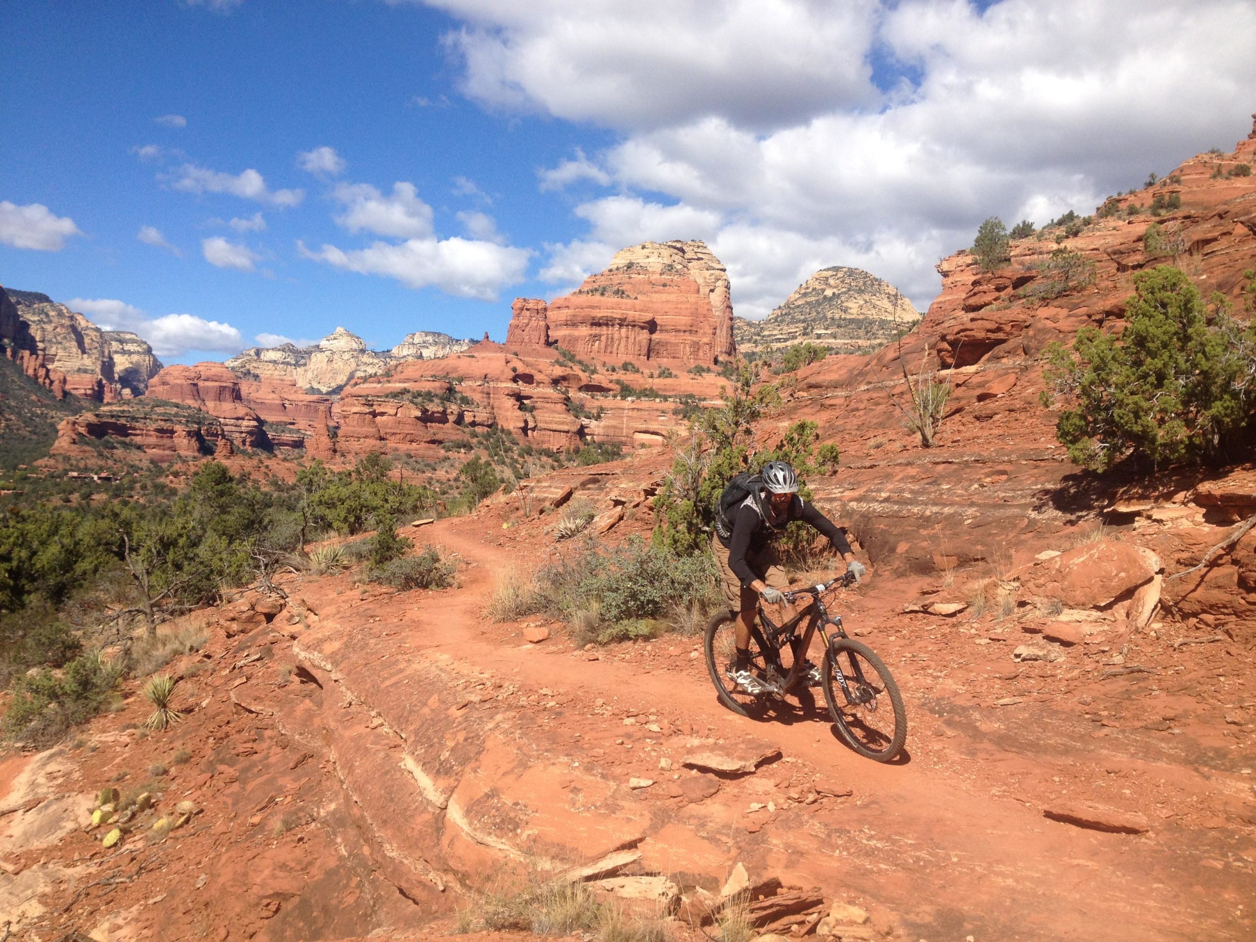 A mountain biker rides along a rocky trail through a desert landscape, surrounded by red rock formations and green shrubs under a partly cloudy sky. Upper Dry Creek Area Trails mountain bike trail.