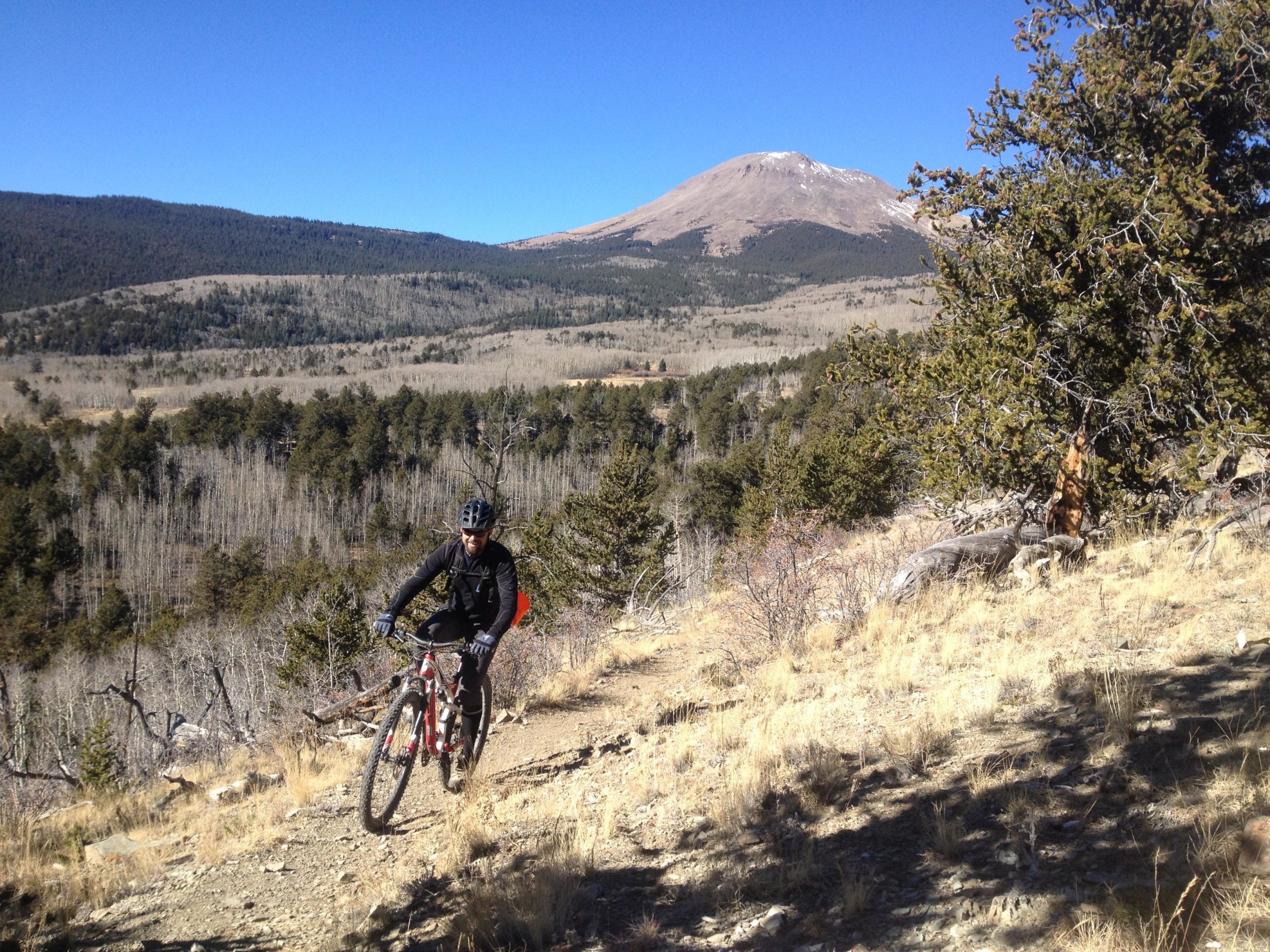 A mountain biker riding uphill on a rocky trail with a scenic view of mountains and trees in the background under a clear blue sky. Salt Creek Trail mountain bike trail.