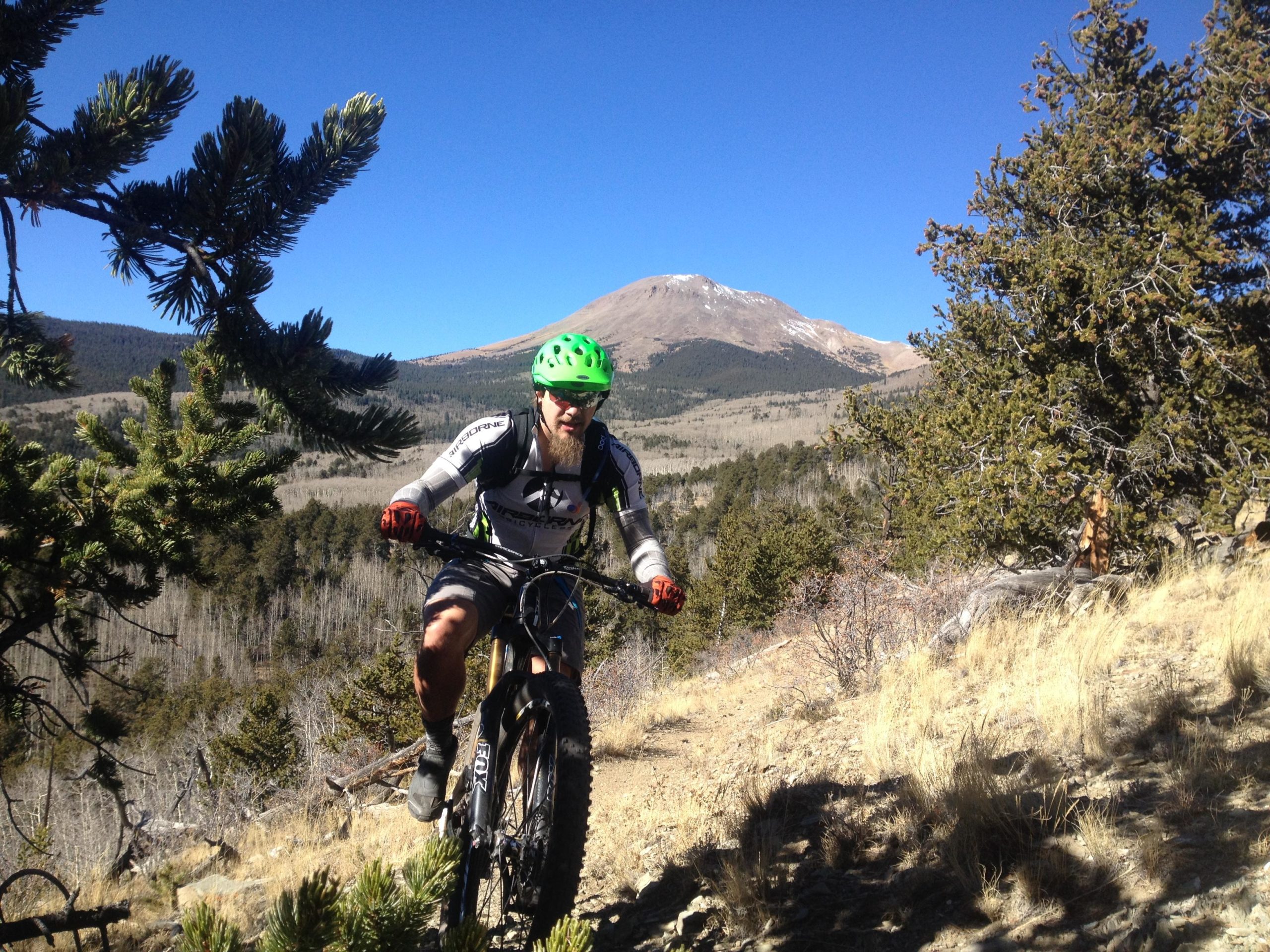 A mountain biker navigating a rocky trail surrounded by trees and mountains under a clear blue sky. The biker is wearing a green helmet and protective gear while riding on a fat bike, with a snow-capped peak visible in the background. Salt Creek Trail mountain bike trail.
