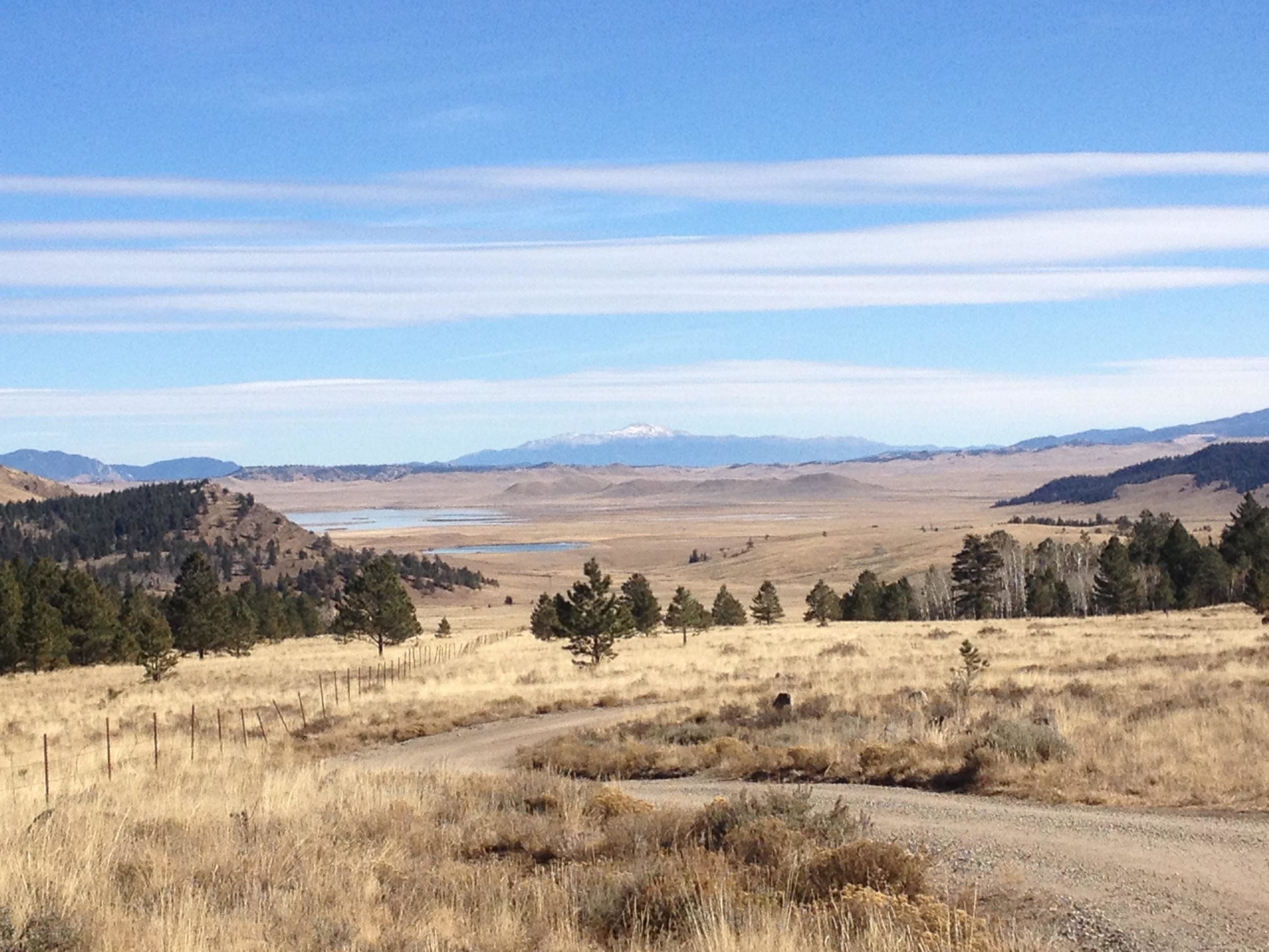 A scenic view of a landscape featuring rolling hills, sparse vegetation, and a clear blue sky. In the foreground, there are patches of dry grass and a winding dirt road bordered by a fence. In the distance, a body of water is visible, surrounded by mountains under a partly cloudy sky. The scene conveys a sense of tranquility and the beauty of nature. Salt Creek Trail mountain bike trail.