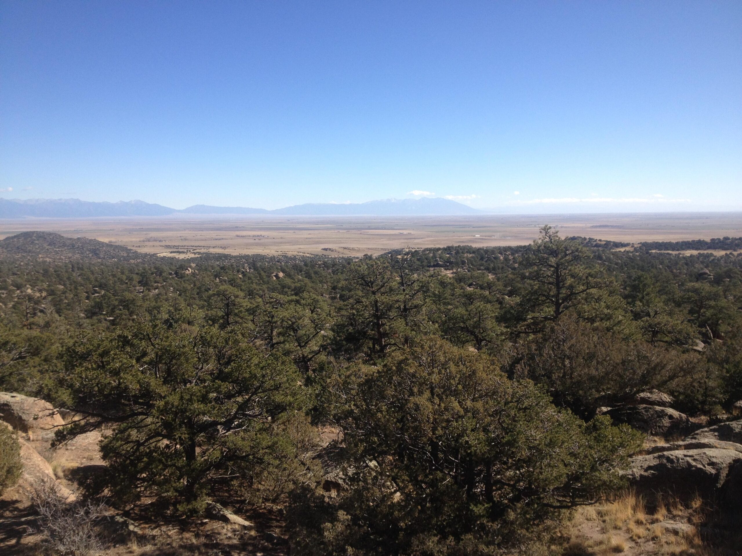 A panoramic view of a valley with sparse vegetation and distant mountains under a clear blue sky. The foreground features green trees and rocky terrain, while the expansive valley stretches into the horizon, leading to misty mountains in the background. Penitente Canyon mountain bike trail.