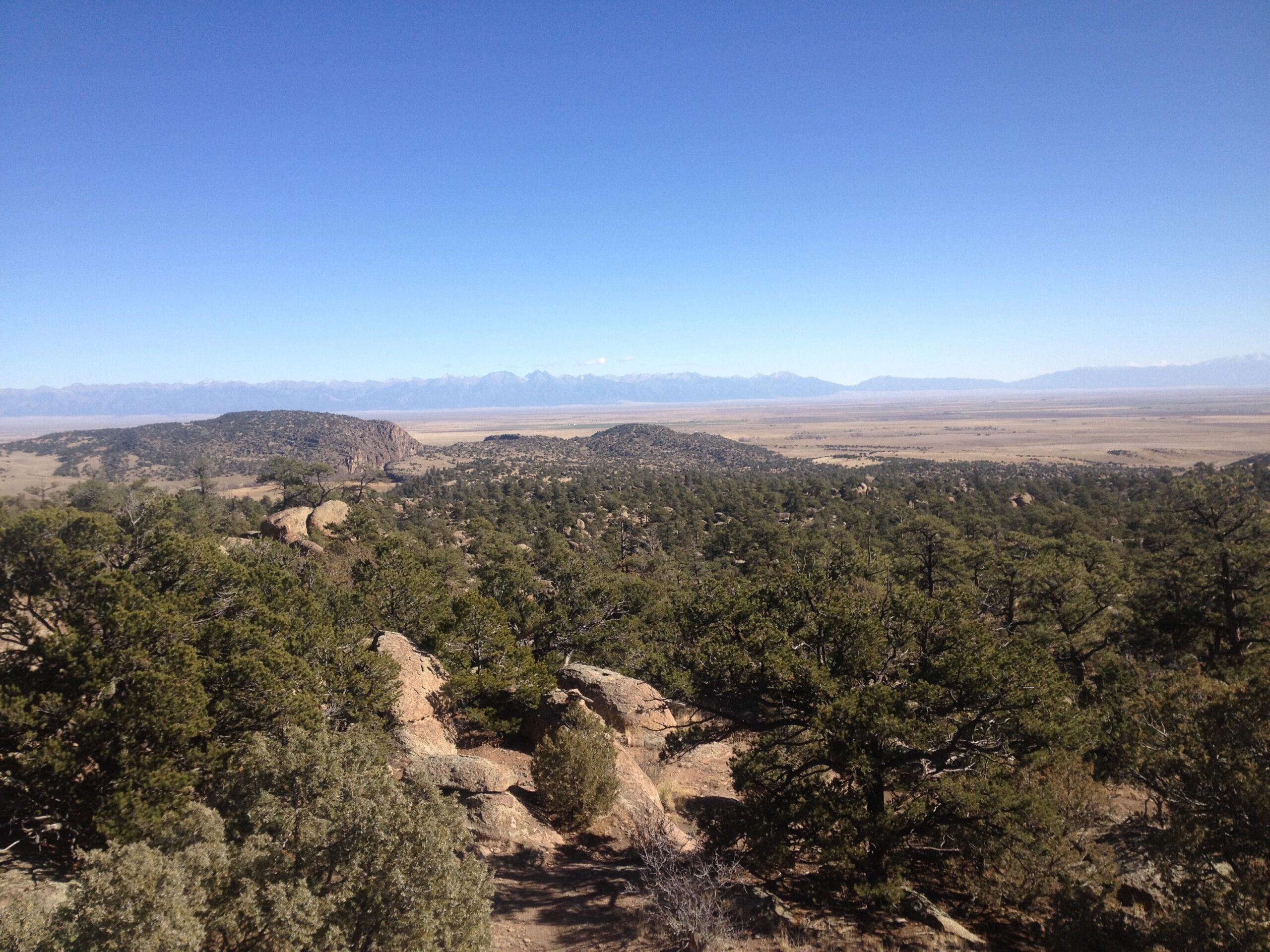 A panoramic view of a mountainous landscape featuring rolling hills, dense patches of green trees, and a vast, open valley under a clear blue sky. The distant mountains are visible against the horizon, creating a scenic backdrop. Penitente Canyon mountain bike trail.