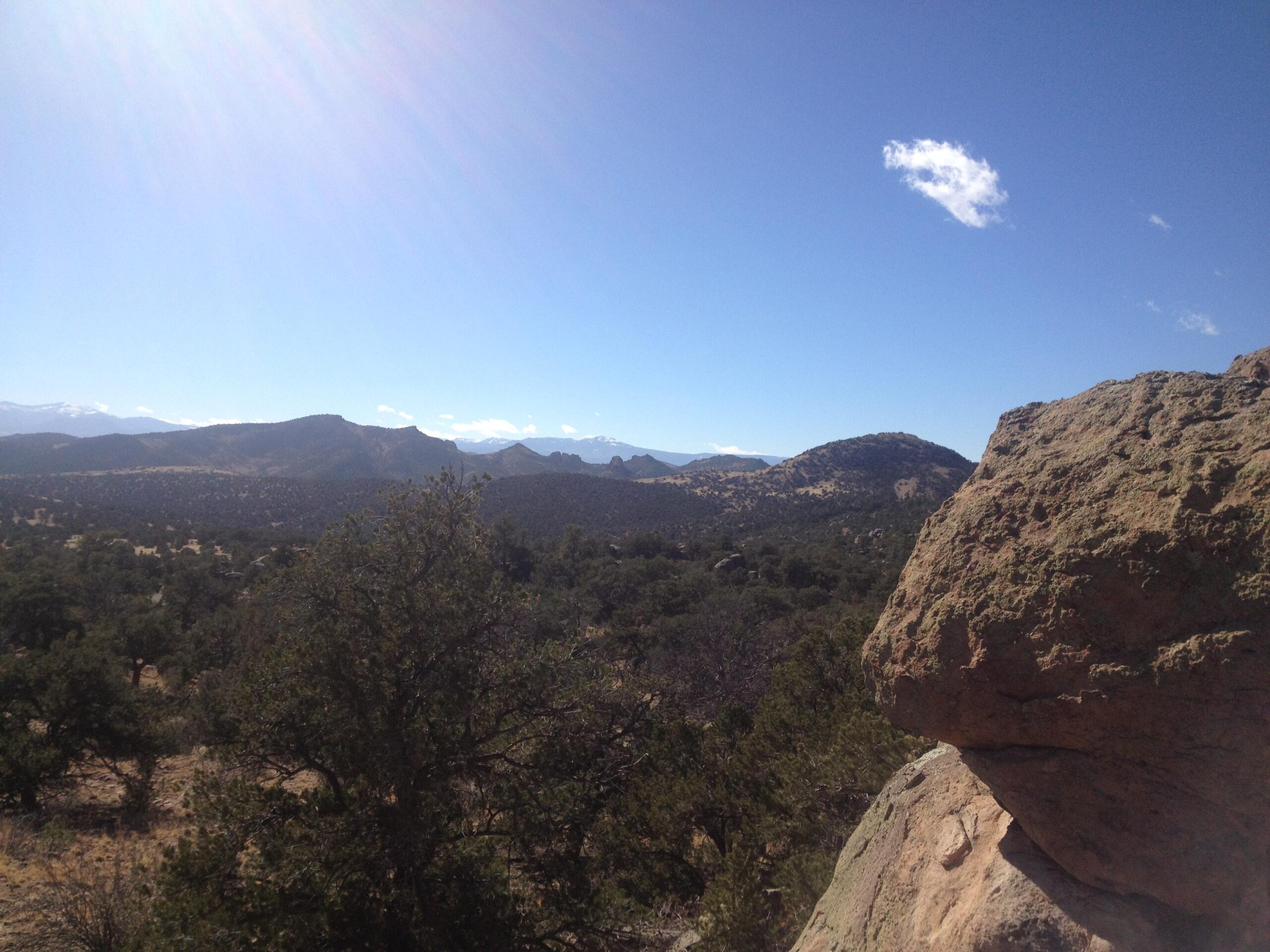 A panoramic view of a mountainous landscape under a clear blue sky, featuring rugged rock formations, scattered trees, and rolling hills in the background. The foreground includes a large rock and patches of vegetation. Penitente Canyon mountain bike trail.