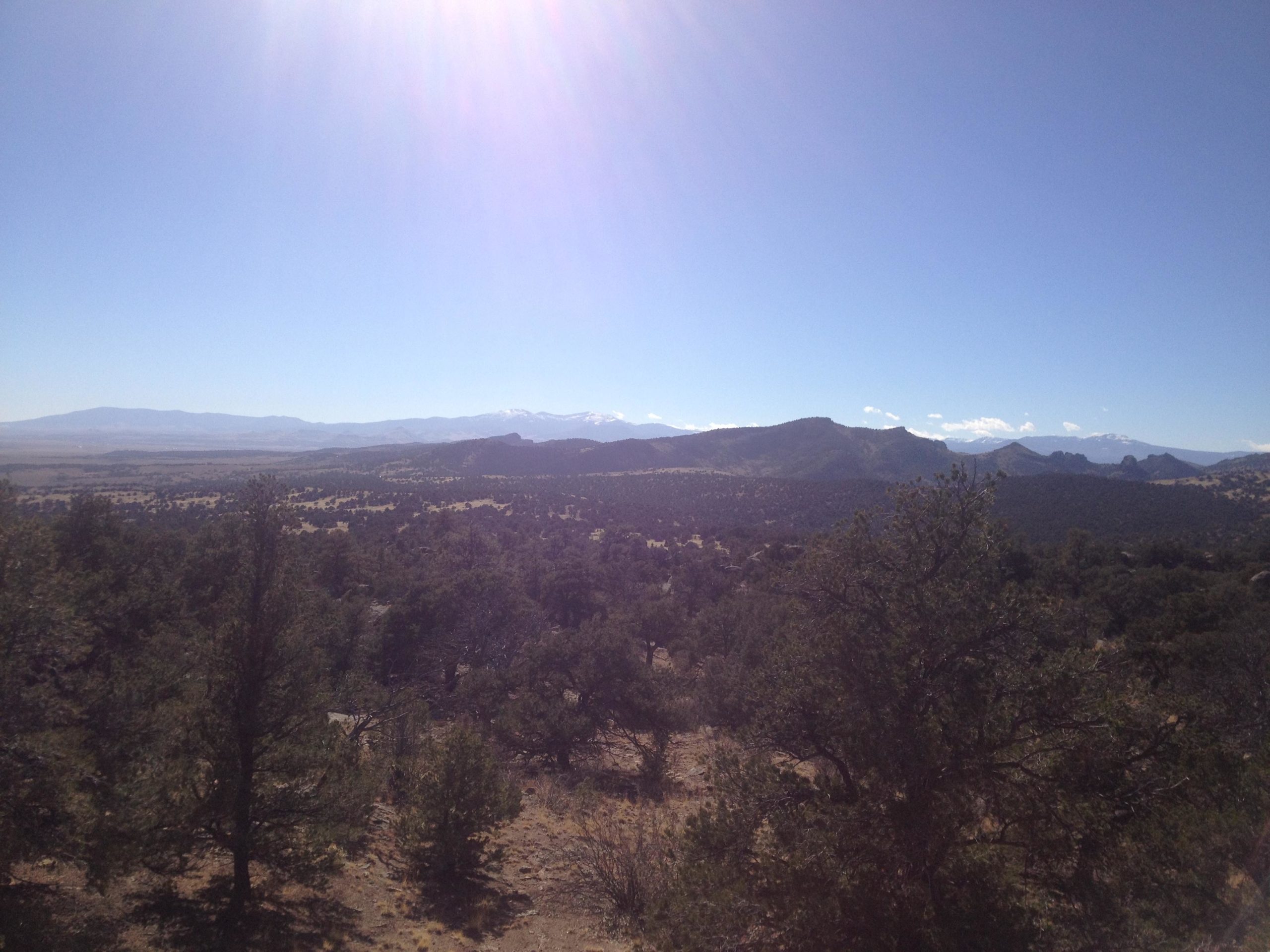 A panoramic view of a mountainous landscape with scattered trees in the foreground, under a clear blue sky. The distant mountains are partially covered with snow, and the sunlight creates a bright glare in the upper left corner. Penitente Canyon mountain bike trail.