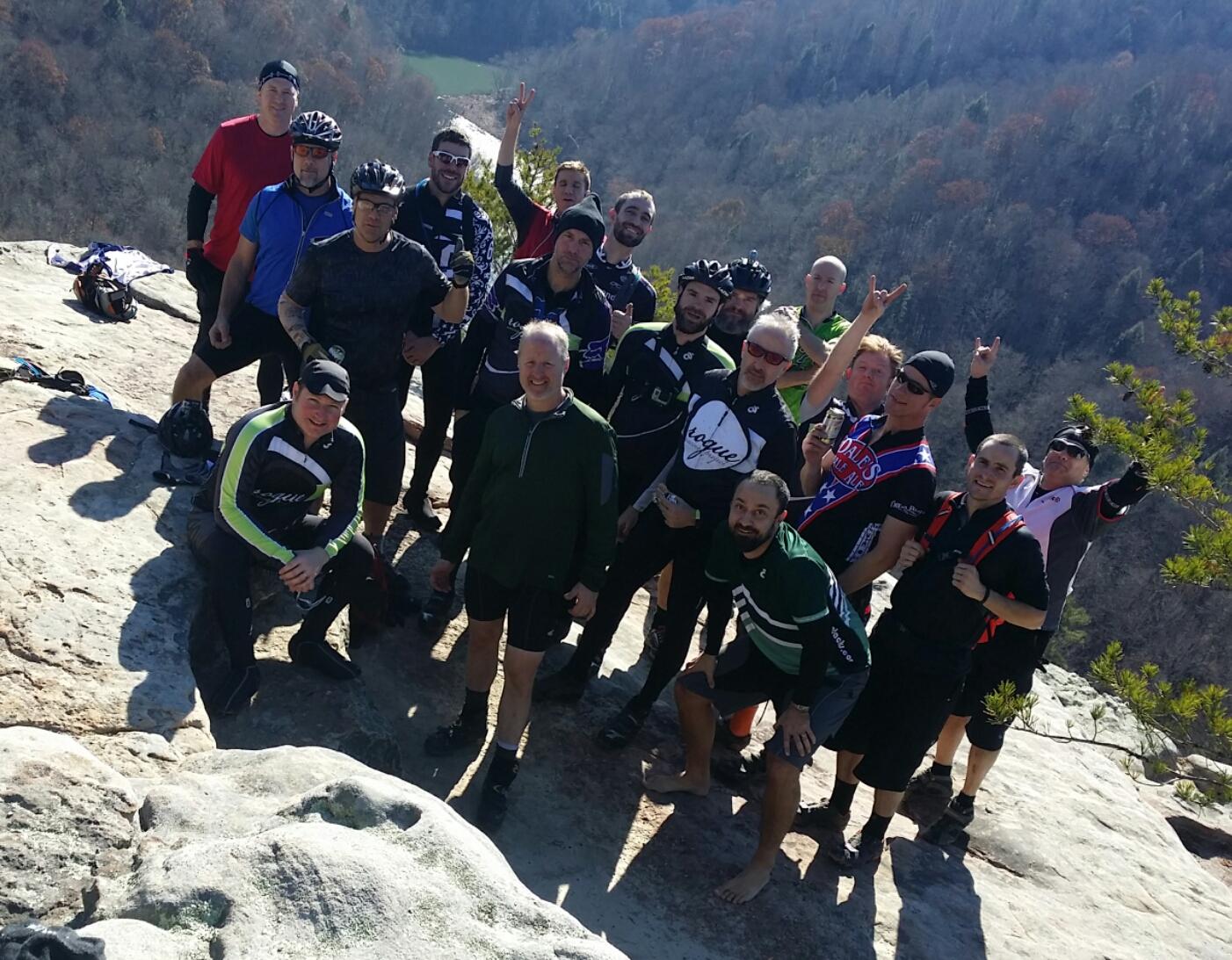 A group of cyclists posing together at a scenic overlook, with trees and hills in the background. They are wearing various cycling jerseys and gear, showing expressions of excitement and camaraderie. Some individuals are making hand gestures, while others are smiling for the camera. Big South Fork mountain bike trail.