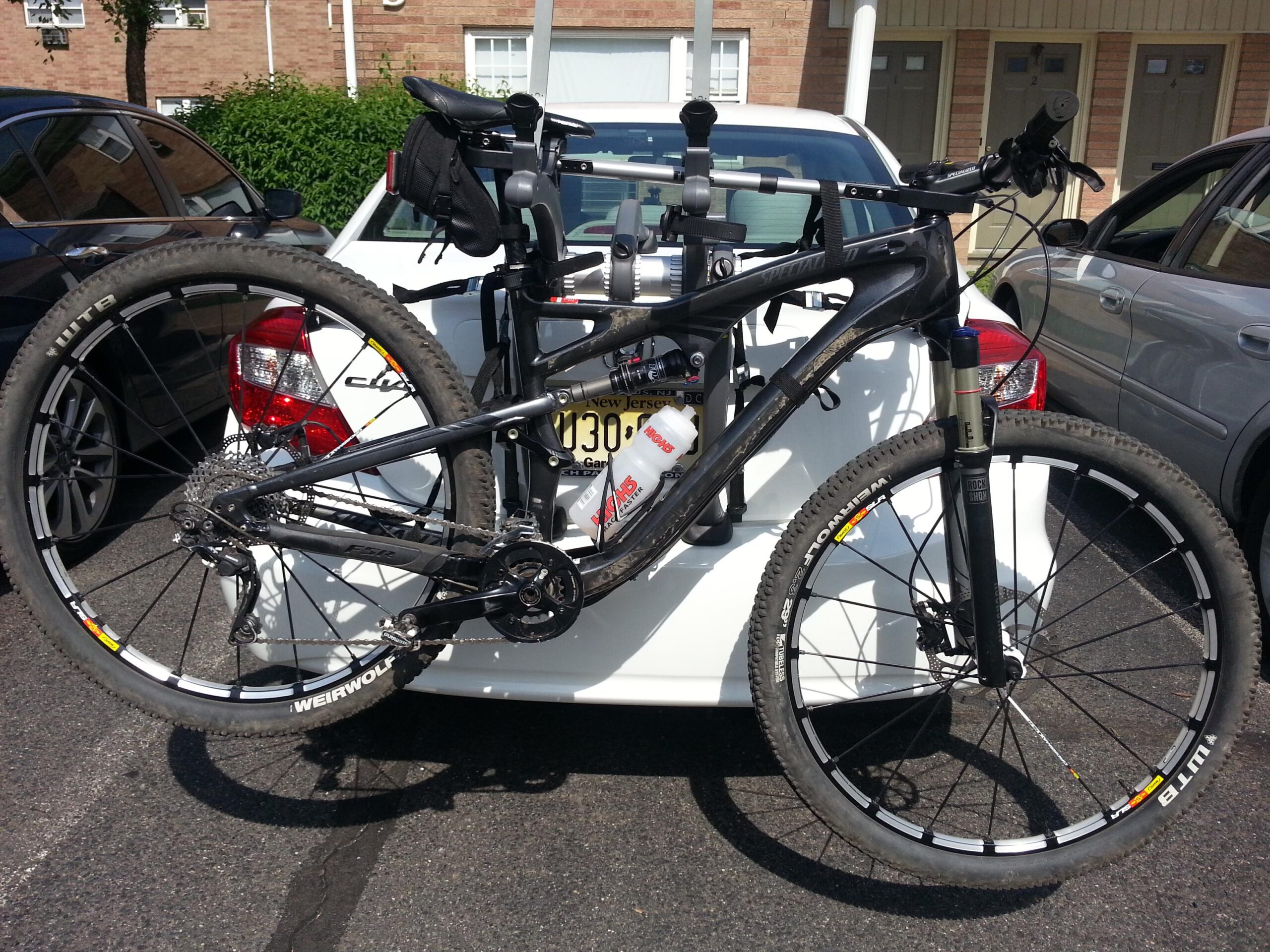 Specialized Camber: A black mountain bike mounted on the back of a white car, with one wheel dangling over the edge. The bike features a water bottle holder and a gear setup with visible components. In the background, there are parked cars and a brick building.