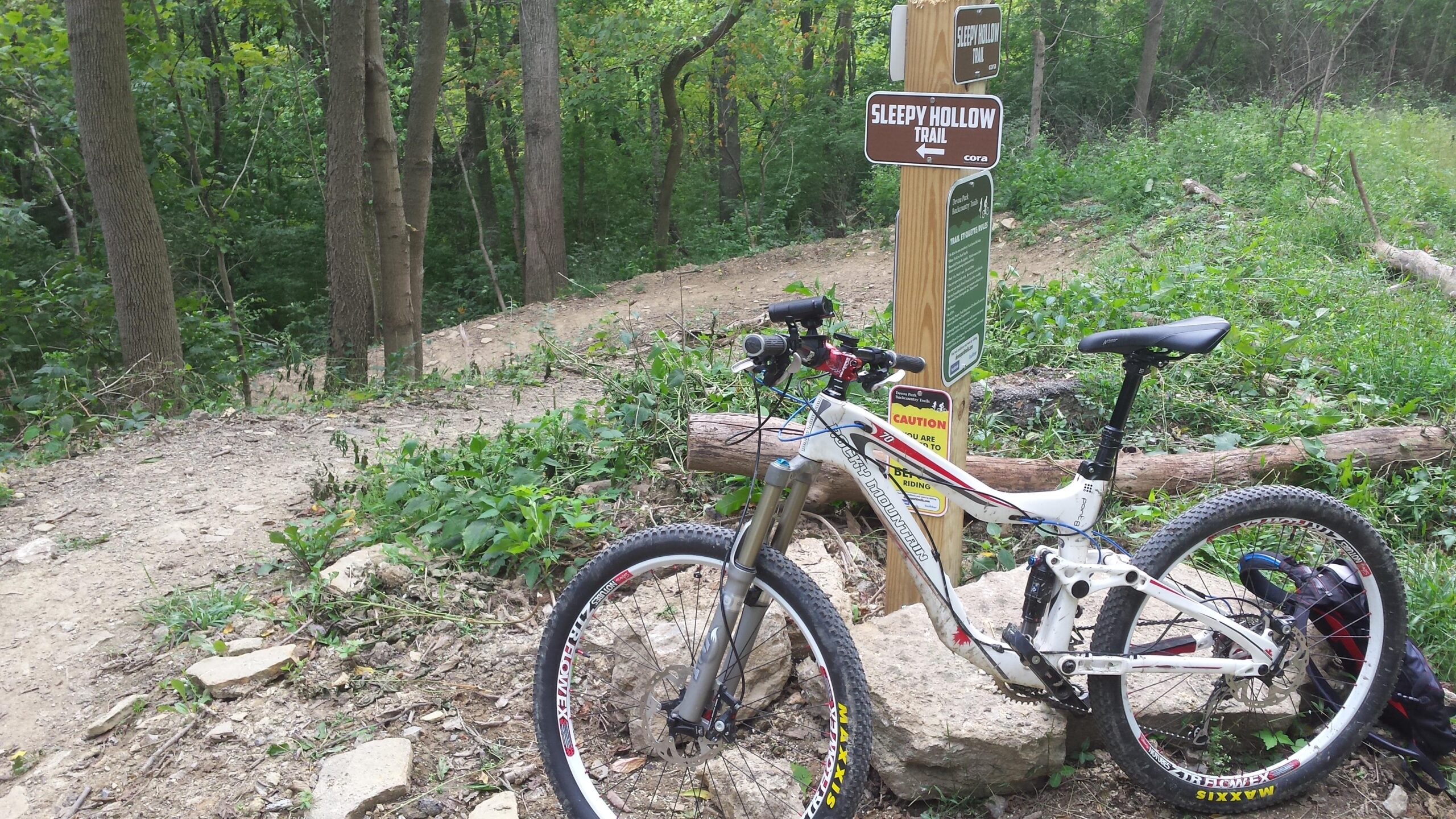 A mountain bike resting against a wooden sign for the Sleepy Hollow Trail, surrounded by lush greenery and trees. The trail is visible in the background, indicating a winding path through the forest. Devou Park mountain bike trail.