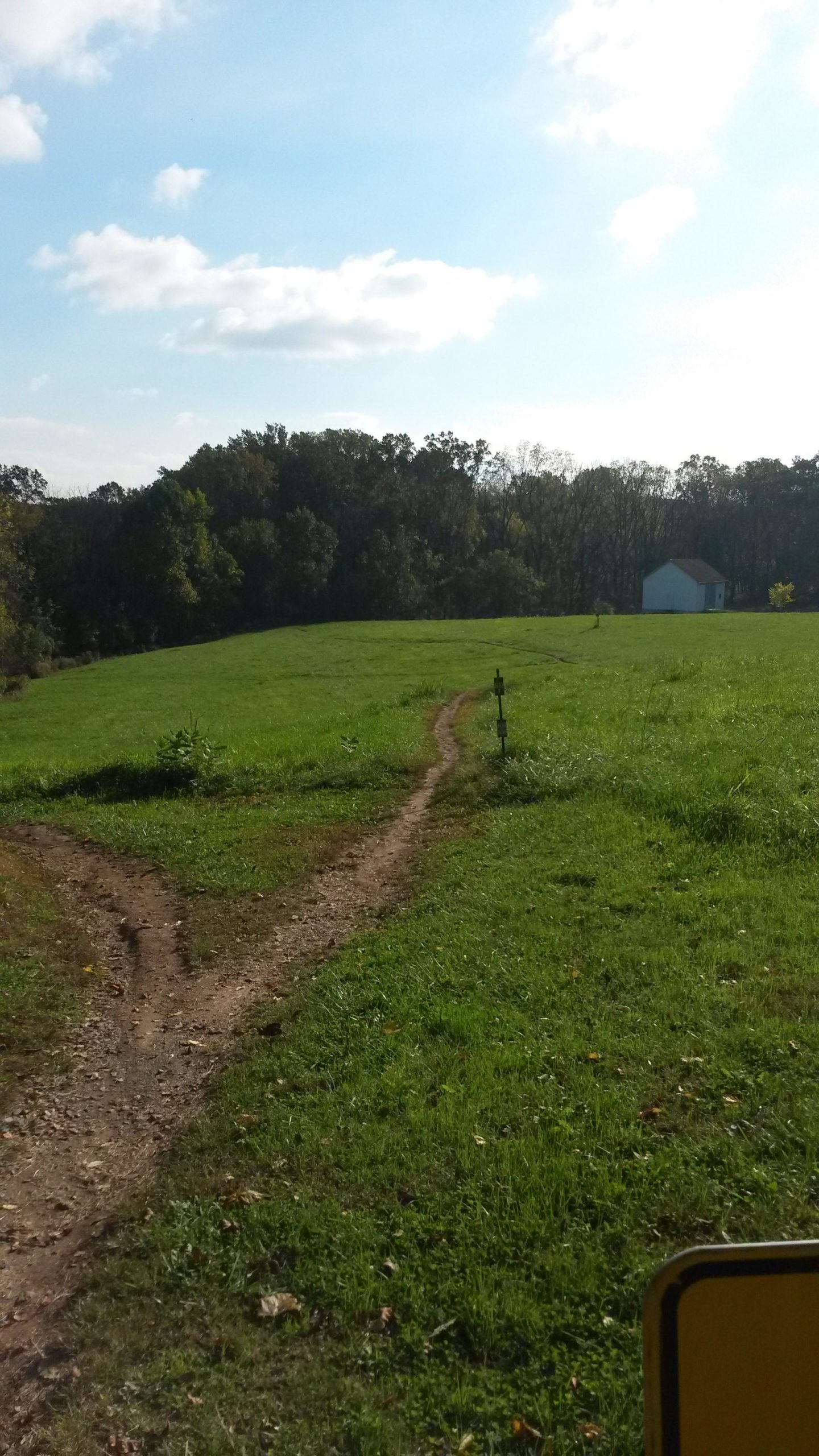 A dirt pathway diverging into two directions through a grassy field, with trees in the background and a small white building to the right under a partly cloudy sky. White Clay Creek mountain bike trail.