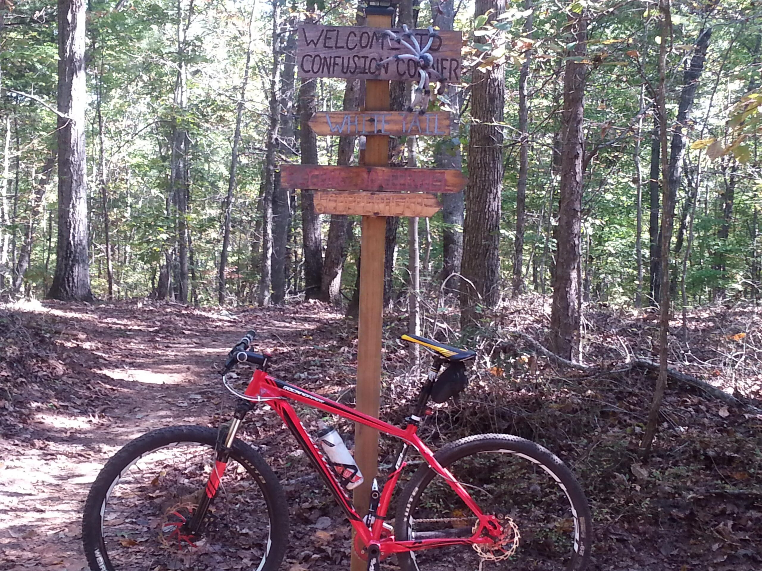 A red mountain bike rests beside a wooden signpost that reads "Welcome to Confusion Corner," with additional signs indicating trail directions. The setting features a lush forest with tall trees and a dirt pathway, suggesting an outdoor recreational area for biking or hiking. Chicopee Woods mountain bike trail.