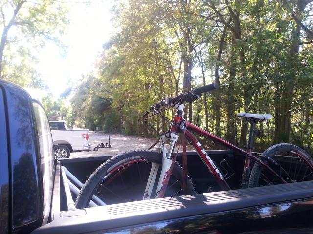 Trek X-Caliber 6: A red mountain bike secured in the bed of a pickup truck, with a grassy path and trees in the background. Another vehicle is visible in the distance along the road.