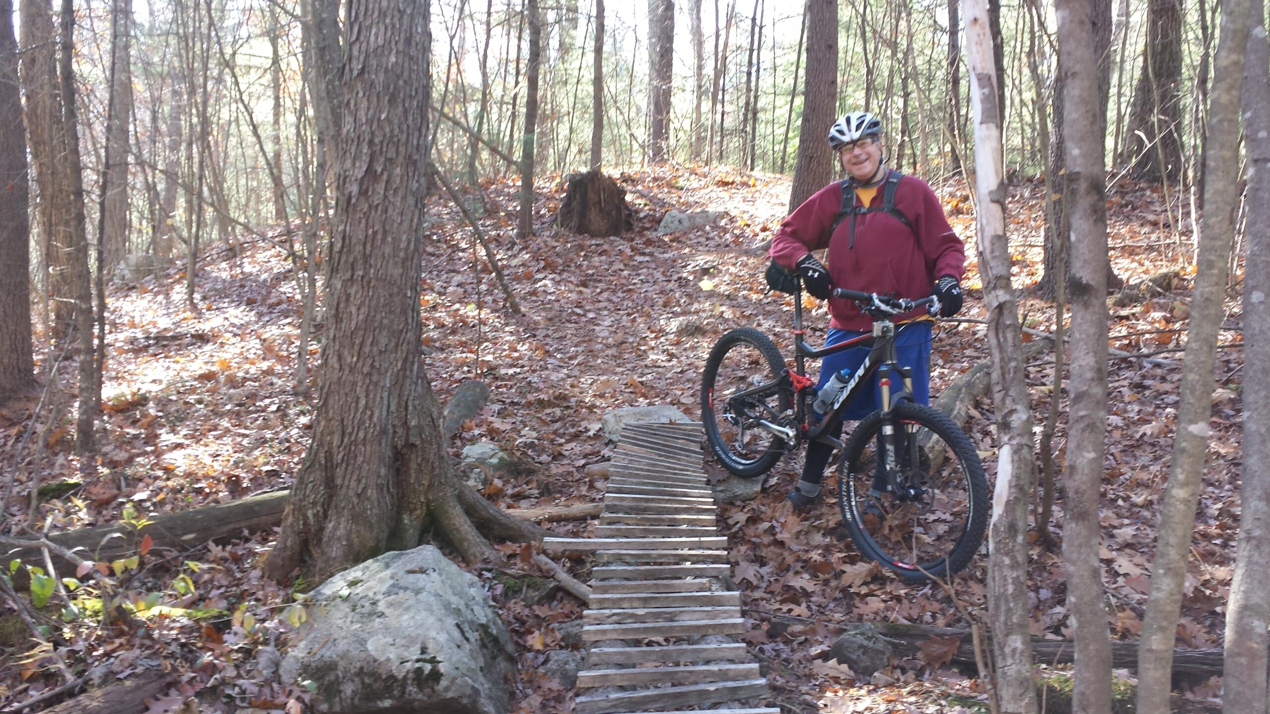 A person wearing a helmet and gloves stands next to a mountain bike on a wooden bridge in a forest. The ground is covered with fallen leaves, and there are trees surrounding the area, creating a scenic and natural backdrop. Musquash Conservation Area mountain bike trail.