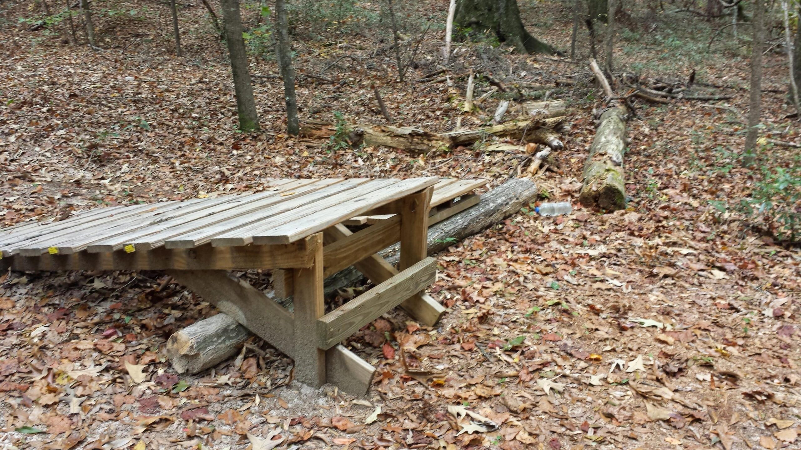 A wooden bridge partially constructed over a forest floor covered with fallen leaves, surrounded by trees and scattered branches. A fallen log is visible to the right, and a plastic bottle is partially obscured among the leaves. Fort Benning MTB Trail mountain bike trail.