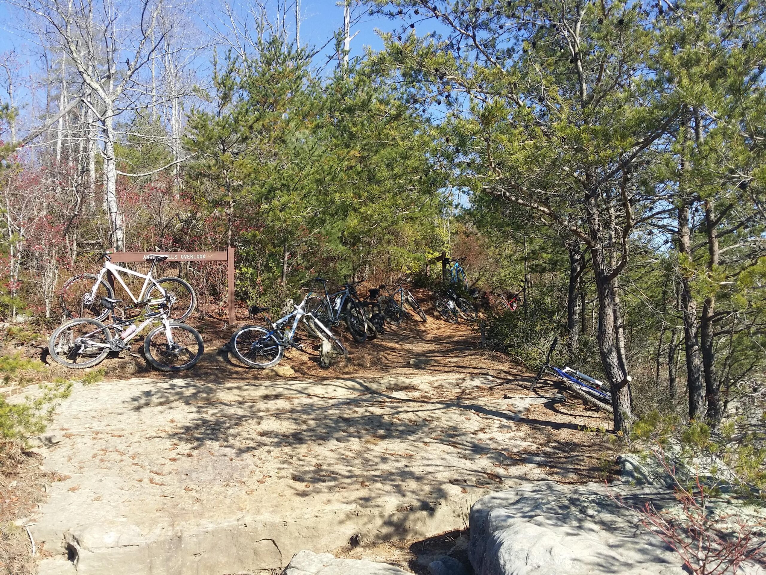 Bicycles are parked along a rocky path in a wooded area, with green trees and some sparse foliage visible. A sign labeled "Overlook" stands nearby, indicating a viewpoint. The scene is bright and sunny, showcasing a clear blue sky. Big South Fork mountain bike trail.