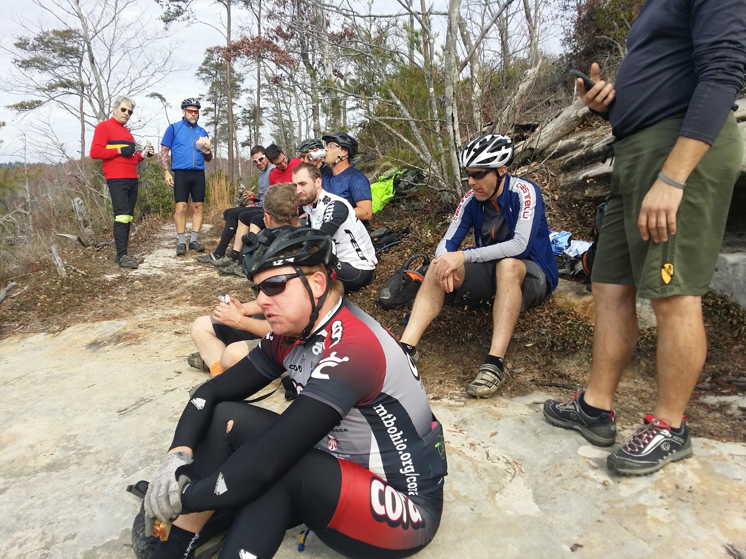 A group of mountain bikers resting on a rocky surface in a wooded area. Some riders are sitting on the ground, while others stand nearby, enjoying snacks and drinks. They are dressed in cycling gear, including helmets and padded shorts. The environment features sparse trees and a mix of earth tones, indicating an outdoor trail setting. John Muir Trail along Chestnut RIdge mountain bike trail.