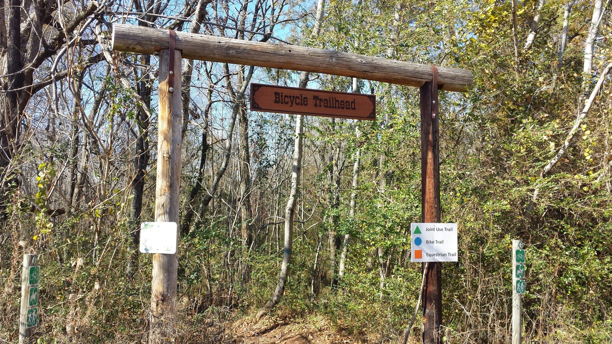 A wooden sign marking the entrance to a bicycle trailhead, surrounded by trees and greenery. The sign reads "Bicycle Trailhead," with additional signage indicating adjacent joint use, bike, and equestrian trails. Heritage Park mountain bike trail.