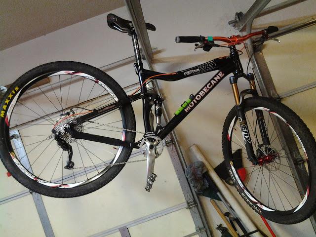 A black mountain bike with red and green accents, hanging upside down in a garage. The bike features thick tires, a visible gear mechanism, and the brand name "Motobecane" displayed prominently on the frame. In the background, there are tools and other items typical of a garage setting.