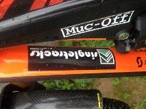 Close-up view of a bicycle frame featuring two stickers: one labeled "Muc-Off" and another with the text "IngaLetercuts.com," both positioned on an orange-colored frame. The scene includes a blurred background of greenery. Sorel Point to L'Etacq mountain bike trail.