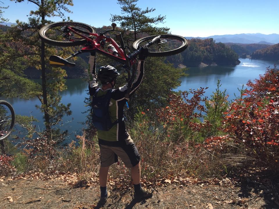 A cyclist wearing a helmet and a backpack raises a mountain bike above their head, standing on a hillside overlooking a serene lake surrounded by autumn foliage and distant mountains under a clear blue sky. Tsali Thompson Loop mountain bike trail.