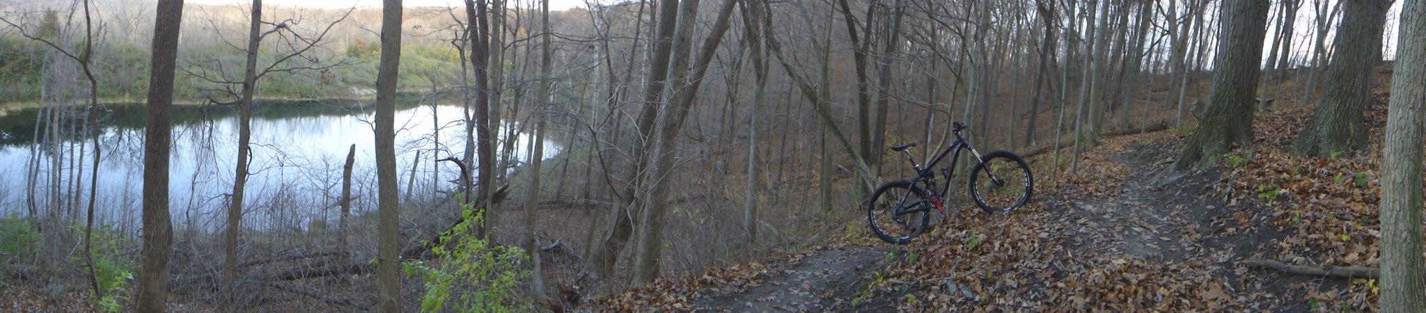 A panoramic view of a wooded area beside a calm lake. The scene features bare trees and fallen leaves on the ground, with a mountain bike leaning against a tree near a dirt path that winds along the water's edge. The setting conveys a tranquil, natural environment, perfect for outdoor activities like biking or hiking. Kickapoo mountain bike trail.