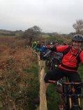 A group of cyclists poses by a wooden fence along a rural path, with a cloudy sky in the background. One cyclist in a red jacket is smiling and leaning against the fence, while several bikes are lined up behind them. Sorel Point to L'Etacq mountain bike trail.