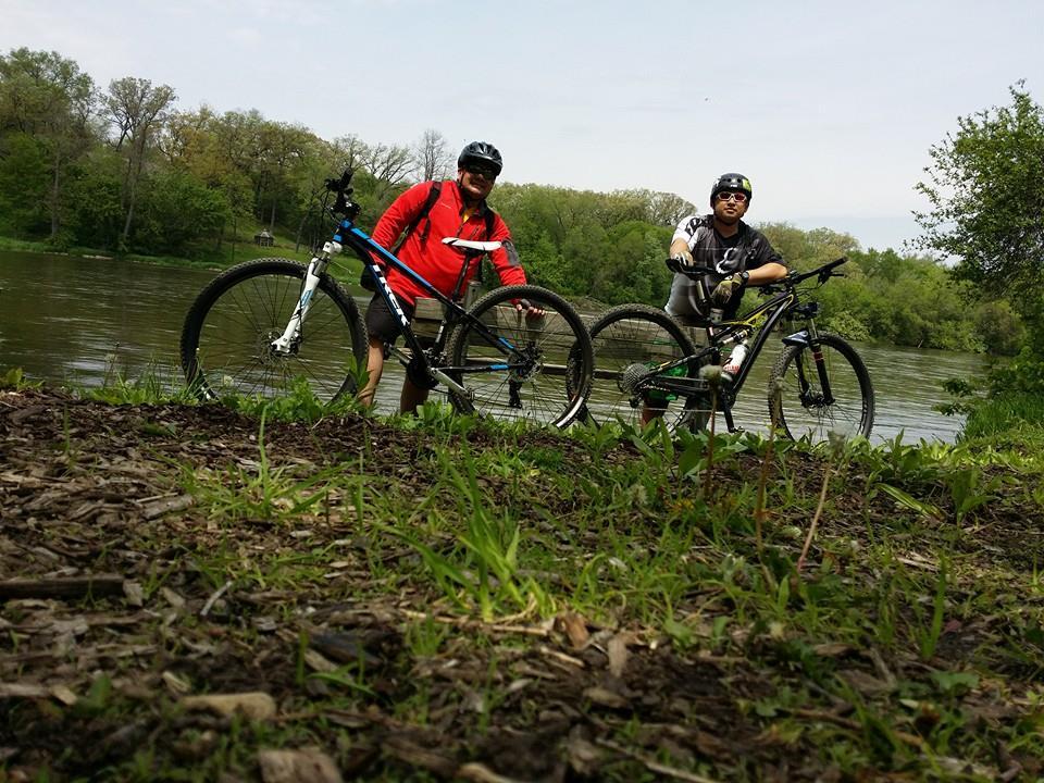 Two mountain bikers posing by a riverbank, surrounded by greenery. One is wearing a red jacket and the other a black cycling jersey, both with helmets on. Their bikes stand next to them as they enjoy the outdoor scenery. Saw Wee Kee Park mountain bike trail.