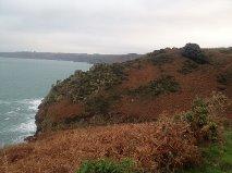 A scenic view of a coastal cliff with rocky outcrops and vegetation, overlooking a calm sea under a cloudy sky. Sorel Point to L'Etacq mountain bike trail.