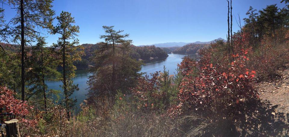 A panoramic view of a serene lake surrounded by autumn foliage, featuring vibrant red and orange leaves on nearby trees. The clear blue sky and distant mountains create a picturesque landscape, reflecting the beauty of nature in the fall. Tsali Thompson Loop mountain bike trail.