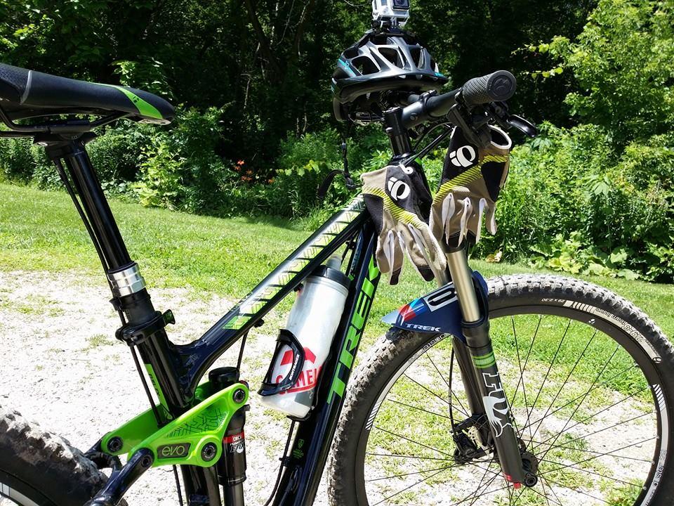 Trek Fuel EX 7 29: A close-up view of a mountain bike leaning against a grassy area. The bike features a black frame with green accents, a water bottle mounted on the side, and a helmet placed on the handlebars. A pair of gloves hangs from the handlebars. The background consists of lush greenery, enhancing the outdoor activity atmosphere.