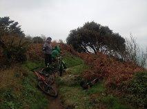 Two cyclists pause on a narrow, grassy trail surrounded by greenery and bushes. One cyclist is wearing a gray jacket, while the other is dressed in green. Mountain bikes are parked nearby, and the scene is set against an overcast sky. Sorel Point to L'Etacq mountain bike trail.