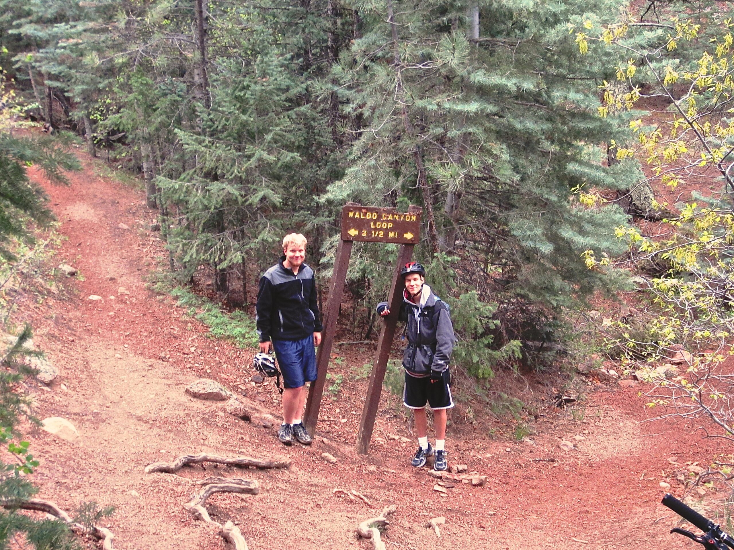 Two young men standing next to a trail sign labeled "Waldo Canyon Loop 1/2 MI" in a forested area. The trail, marked by reddish soil and surrounded by trees, leads into the distance. One of the men is holding a bicycle helmet, while both are dressed in casual outdoor attire. Waldo Canyon mountain bike trail.