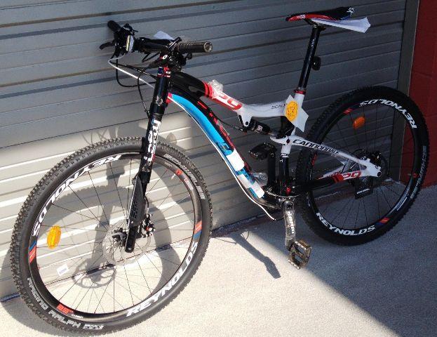 Cannondale Scalpel 29er 1: A black and blue mountain bike with red and white accents, featuring a lightweight frame, front suspension, and knobby tires, positioned next to a garage door. The bike has a price tag still attached and is resting on a flat surface.