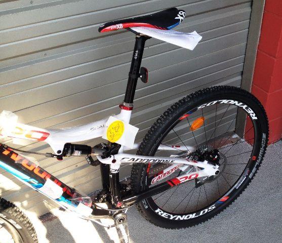 Cannondale Scalpel 29er 1: A close-up view of a Cannondale mountain bike leaning against a metallic storage door. The bike features a sleek white and black frame with colorful accents, a prominent saddle, and Reynolds wheels. A tag can be seen attached to the seat post. The bike's tire has a knobby design, indicating its suitability for off-road riding.