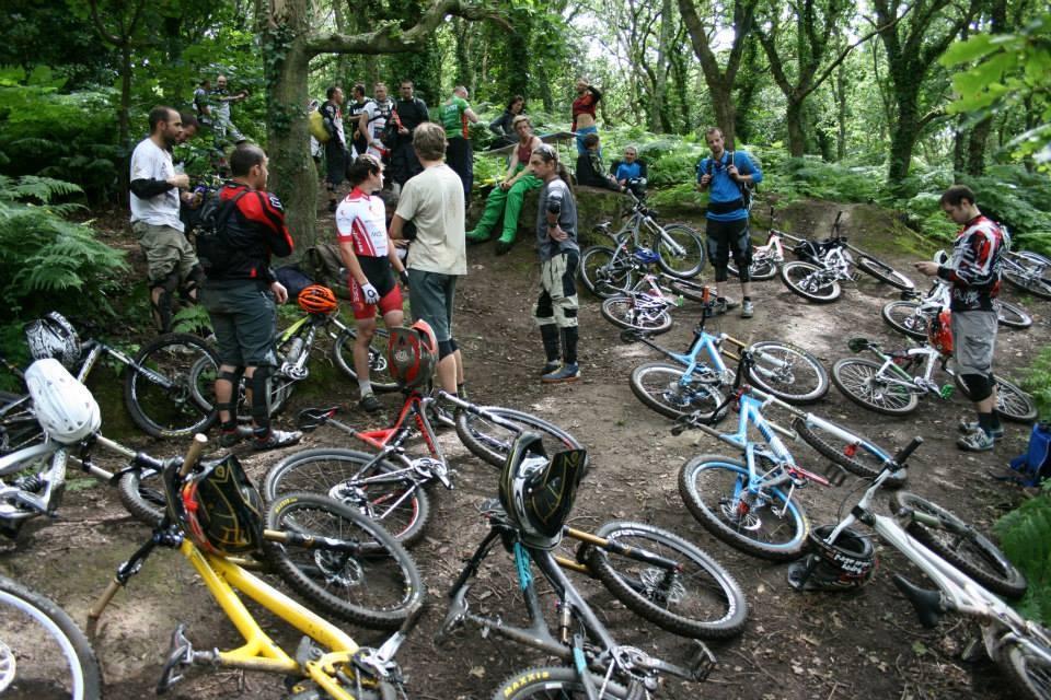 A group of mountain bikers gathered in a wooded area, with several bicycles parked around them. The riders are dressed in biking gear, engaged in conversation, while others are seated on a dirt rise. The scene showcases a vibrant outdoor activity atmosphere, surrounded by lush green foliage.