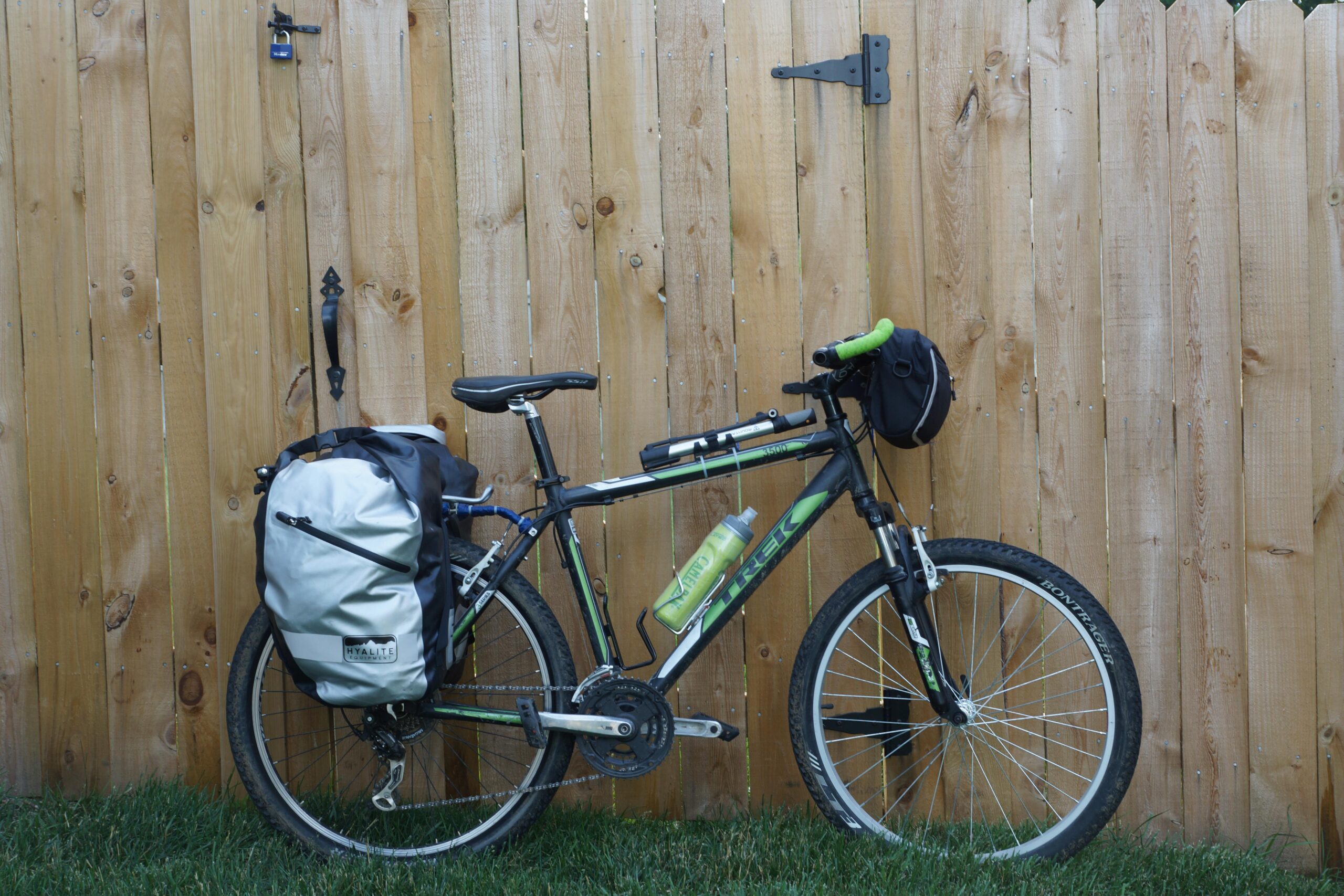 Trek 3500: A bicycle leaning against a wooden fence, featuring a black frame with green accents. It has a large, waterproof bag attached to the rear and a water bottle mounted on the frame. A small black bag is attached to the handlebars. The ground is grassy, and the fence appears to be made of planks.